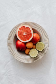 Flat lay of fresh citrus and stone fruits in a textured bowl on white background.