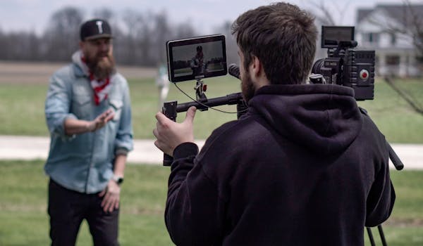 A cameraman films a music video scene outdoors in a field, with professional equipment.