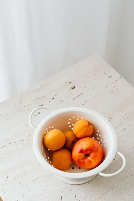 Top view of fresh apricots and a peach in a white strainer on a wooden table.