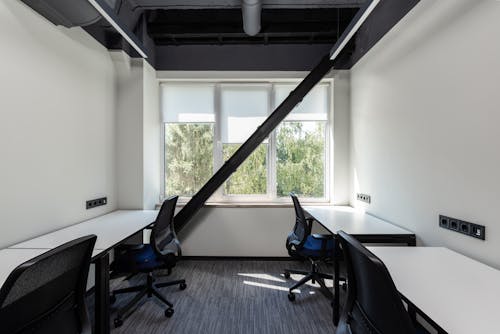 Interior of contemporary light workplace with chairs near tables next to windows with jalousie under ceiling with pipes