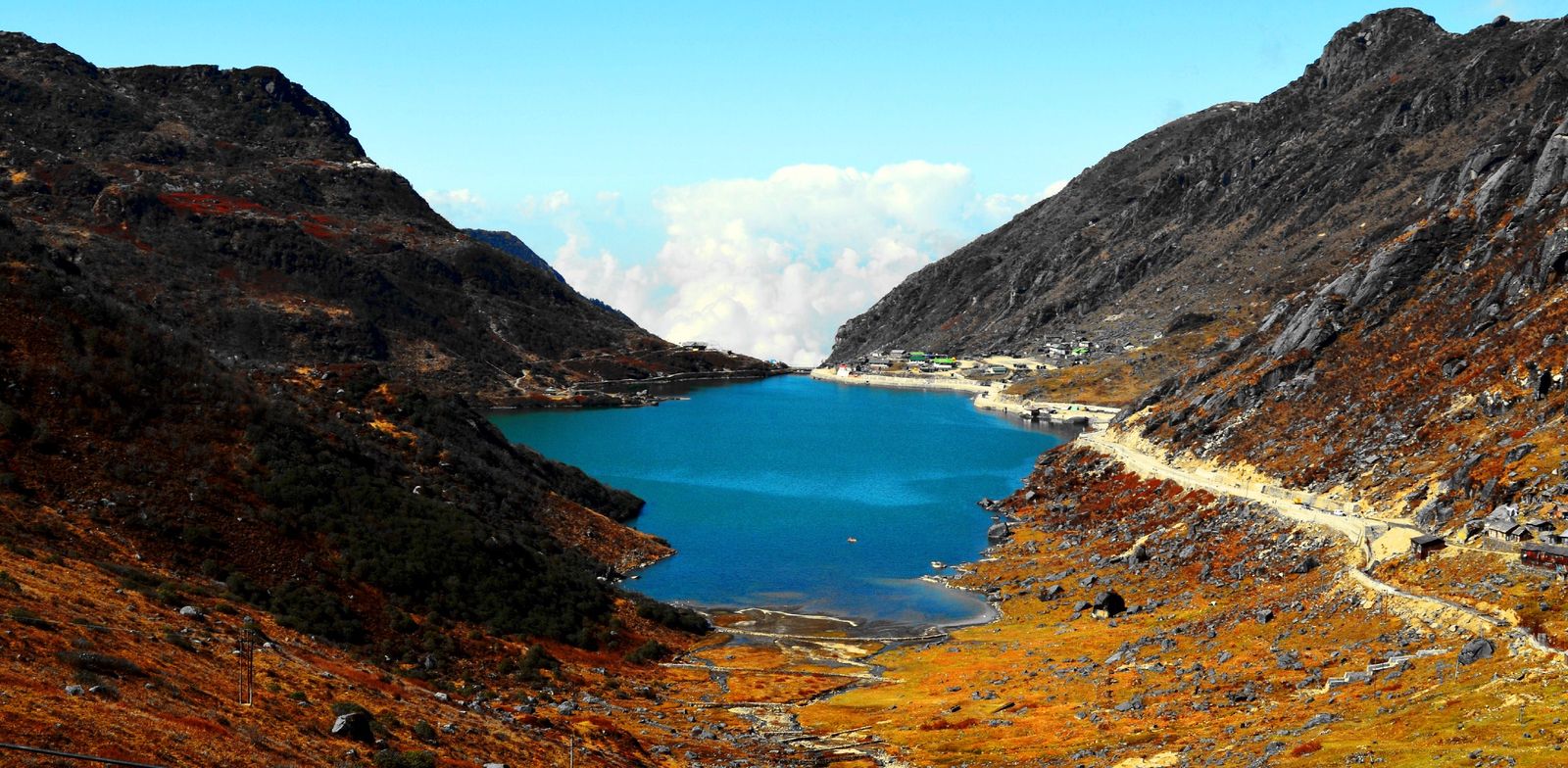 Tsomgo Lake in autumn, blue water between rust brown slopes, the permit road running along its shore