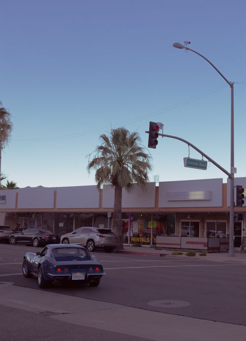 Free Vintage car driving past storefronts on a sunny street corner with palm trees and traffic lights. Stock Photo