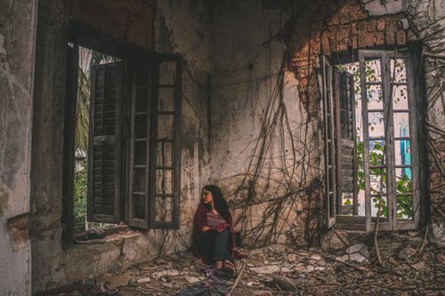 Free A teenager sitting in an abandoned, decaying house with broken windows and growing vines. Stock Photo