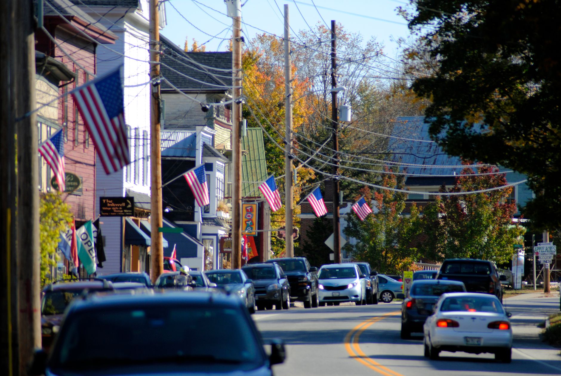 Modern cars driving on road through modern suburban cottages decorated with USA national flags in early autumn