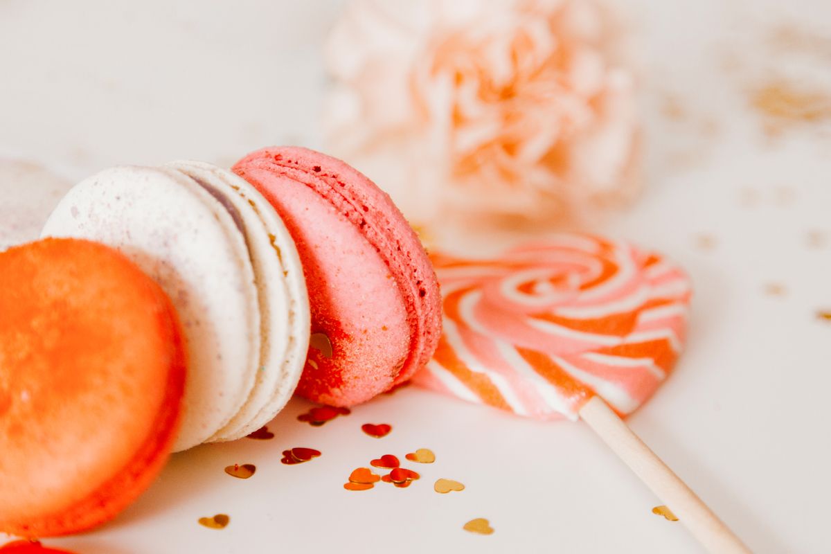 Close-up of vibrant macarons and a lollipop on a white surface, with decorative heart confetti.