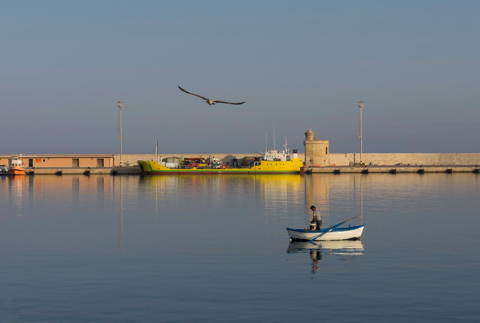A serene scene of a fisherman in a boat at Gallipoli harbor with a bird flying overhead.