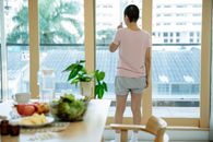 Back view of unrecognizable woman with short hair in casual clothes drinking glass of water and looking through windows while standing at home near table with assorted vegetable and medicines