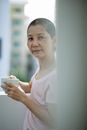 Side view of peaceful young Asian female with dark short hair drinking coffee and looking at camera while standing on terrace in sunny morning