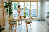 Side view of Asian woman with dark short hair in casual clothes drinking glass of water while standing near table with various fruits and medicines during recovery at home