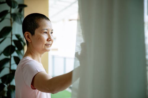 Cheerful ethnic ill female on remedy looking away in window of hospital in daytime