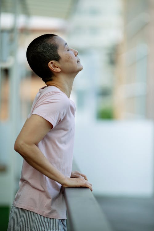 Side view of cheerful ethnic short haired female with eyes closed resting on balcony while leaning on railing in hospital