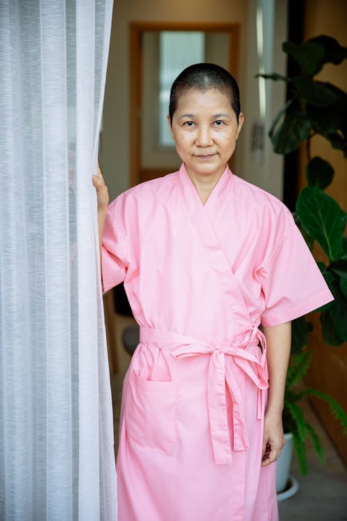 Short haired Asian woman with breast cancer in robe touching curtain