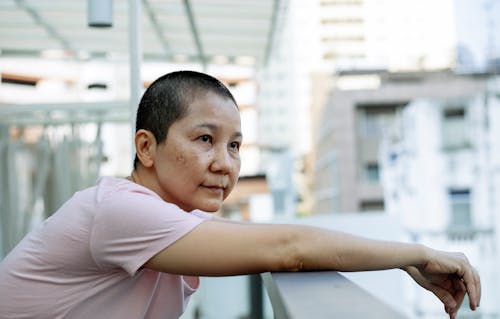 Side view of calm ethnic short haired ill female leaning on railing of terrace of hospital on blurred background