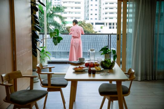 Back view of anonymous female with short hair in pink robe suffering from cancer in comfortable apartment with balcony