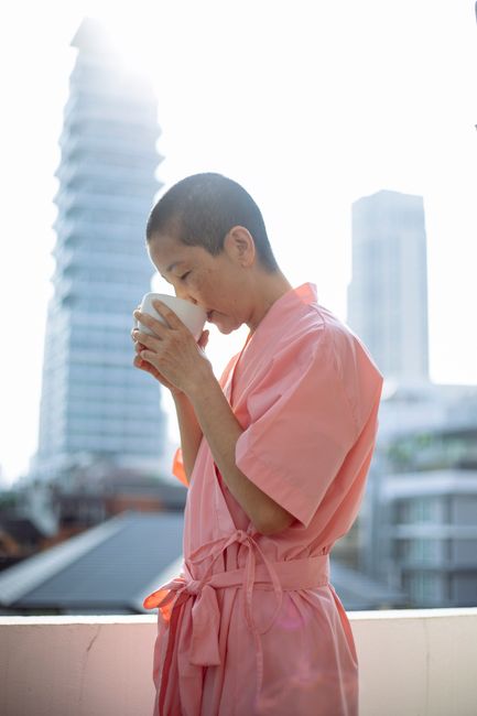 Side view of ethnic slender female in pink robe with hot mug of tea in hospital on blurred background