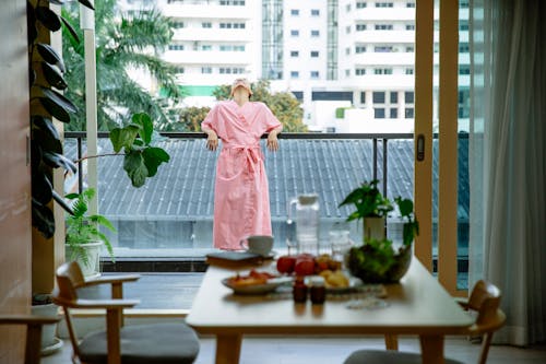 Unrecognizable woman in pink robe standing with raised head on balcony in room with table with fresh food in morning time