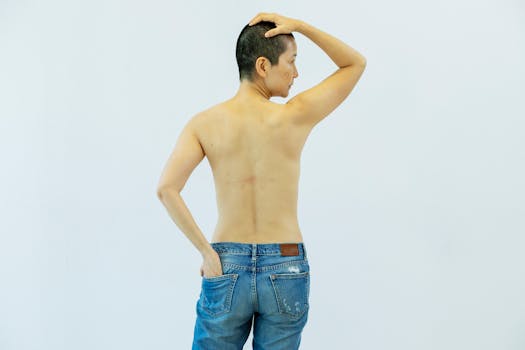 A woman with short hair poses topless in denim jeans against a studio background, showing confidence.