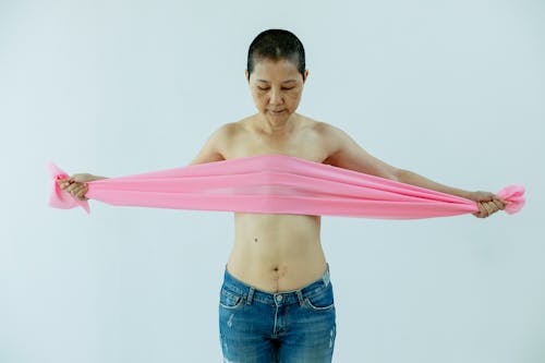 Free Serious topless middle age Asian lady in jeans covering chest with pink scarf after breast removal surgery from cancer wile standing in bright studio on white background Stock Photo