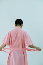Back view of unrecognizable woman in pink bathrobe with belt standing on white background in light studio