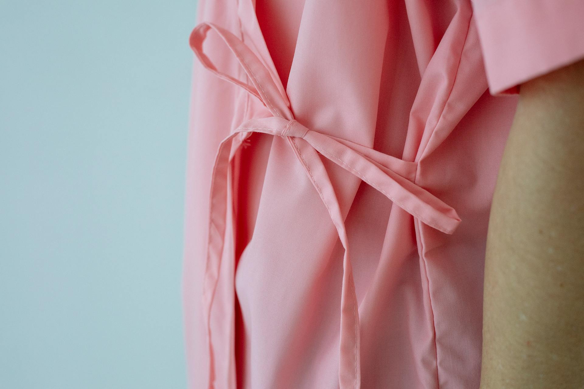 Crop unrecognizable female patient in pink gown with pink ribbon standing against white background