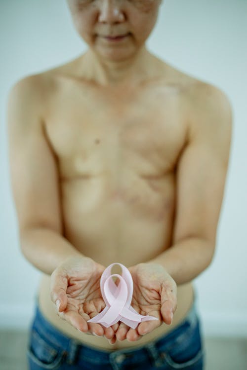 Crop unrecognizable woman with bare torso and scars left after operation holding pink ribbon in support to women with breast cancer on Breast Cancer day