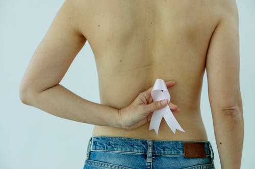 Crop unrecognizable female in blue jeans holding pink ribbon in hand behind back standing against white background