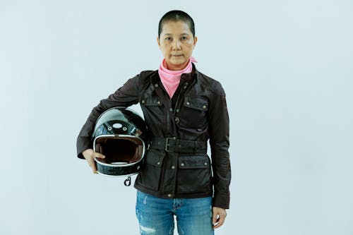 Adult Asian female biker with short hair wearing black leather jacket jeans and pink scarf holding helmet and looking at camera while standing on white background