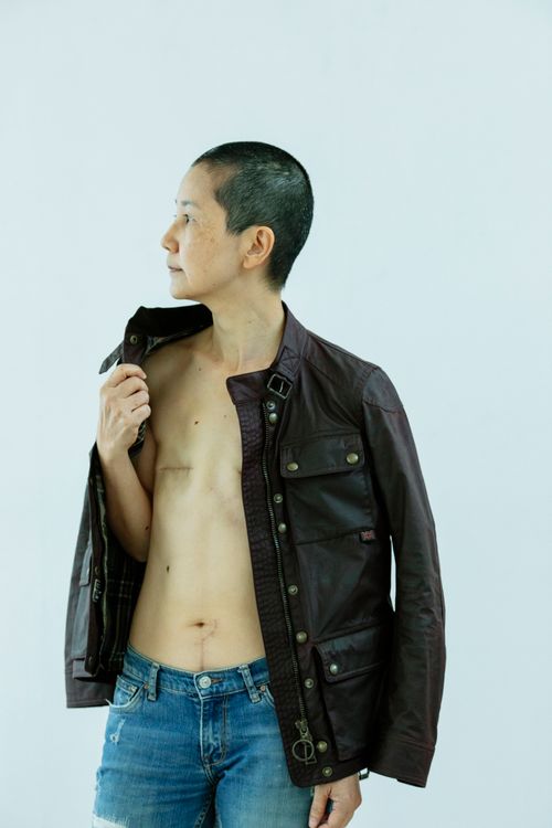 Free Serious middle age Asian female in jeans and jacket with scars on chest after breast removal surgery from breast cancer looking away while standing on white background in light studio Stock Photo