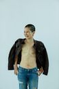 Smiling mature ethnic female in jeans and jacket with scars on chest after breast removal surgery from breast cancer standing in light studio on white background and looking away