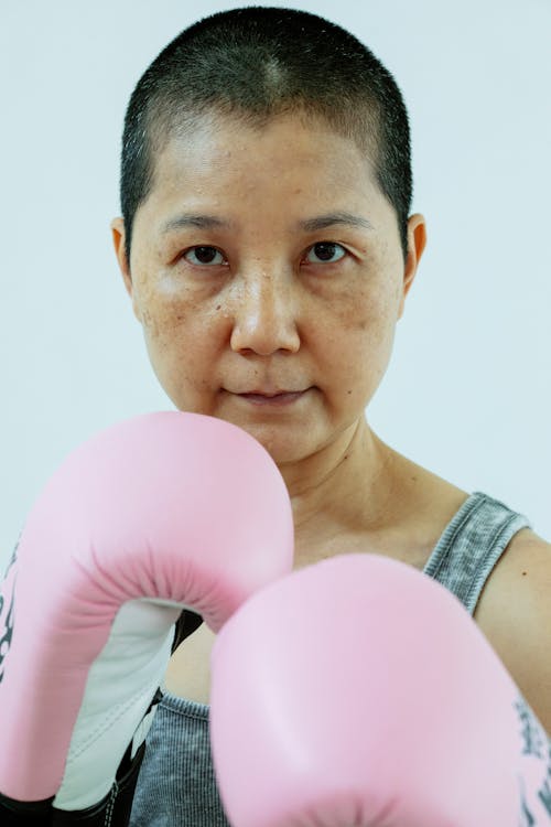 Calm Asian woman with short dark hair in pink boxing gloves looking at camera against gray background in studio