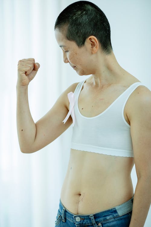 Side view of calm Asian woman with short dark hair in white top with pink ribbon as sign of Breast Cancer Awareness month standing with raised arms in light room