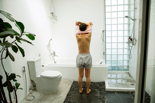 Full length back view of unrecognizable barefoot lady in sleepwear standing and undressing next to bath and toilet with shower in bathroom with green plant