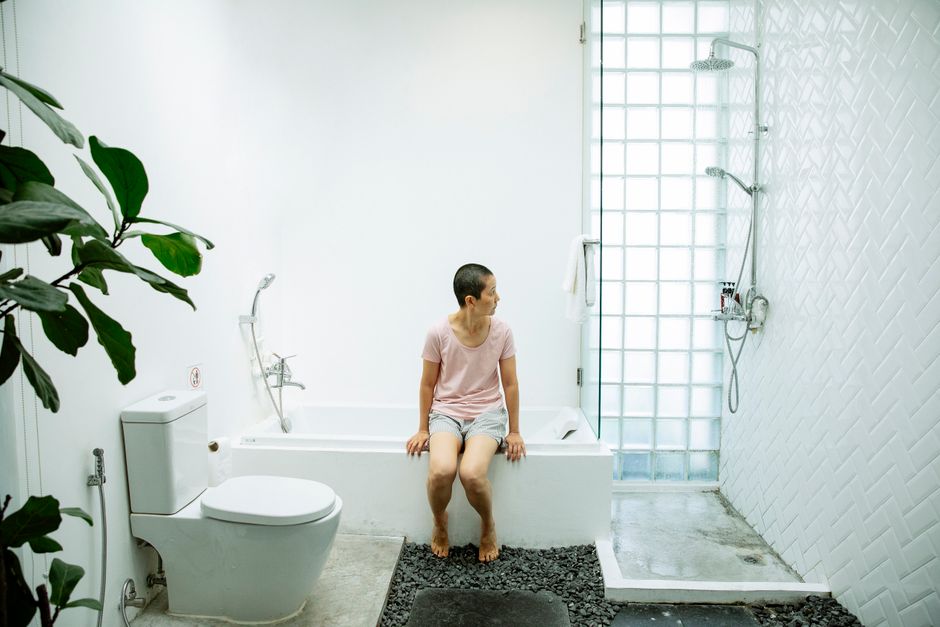 Full body of barefoot female in casual clothes sitting on bathtub next to toilet and shower in bathroom with green plant
