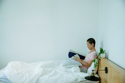 Side view of concentrated lady in sleepwear browsing on cellphone while sitting in bed with pillows under blanket in light bedroom