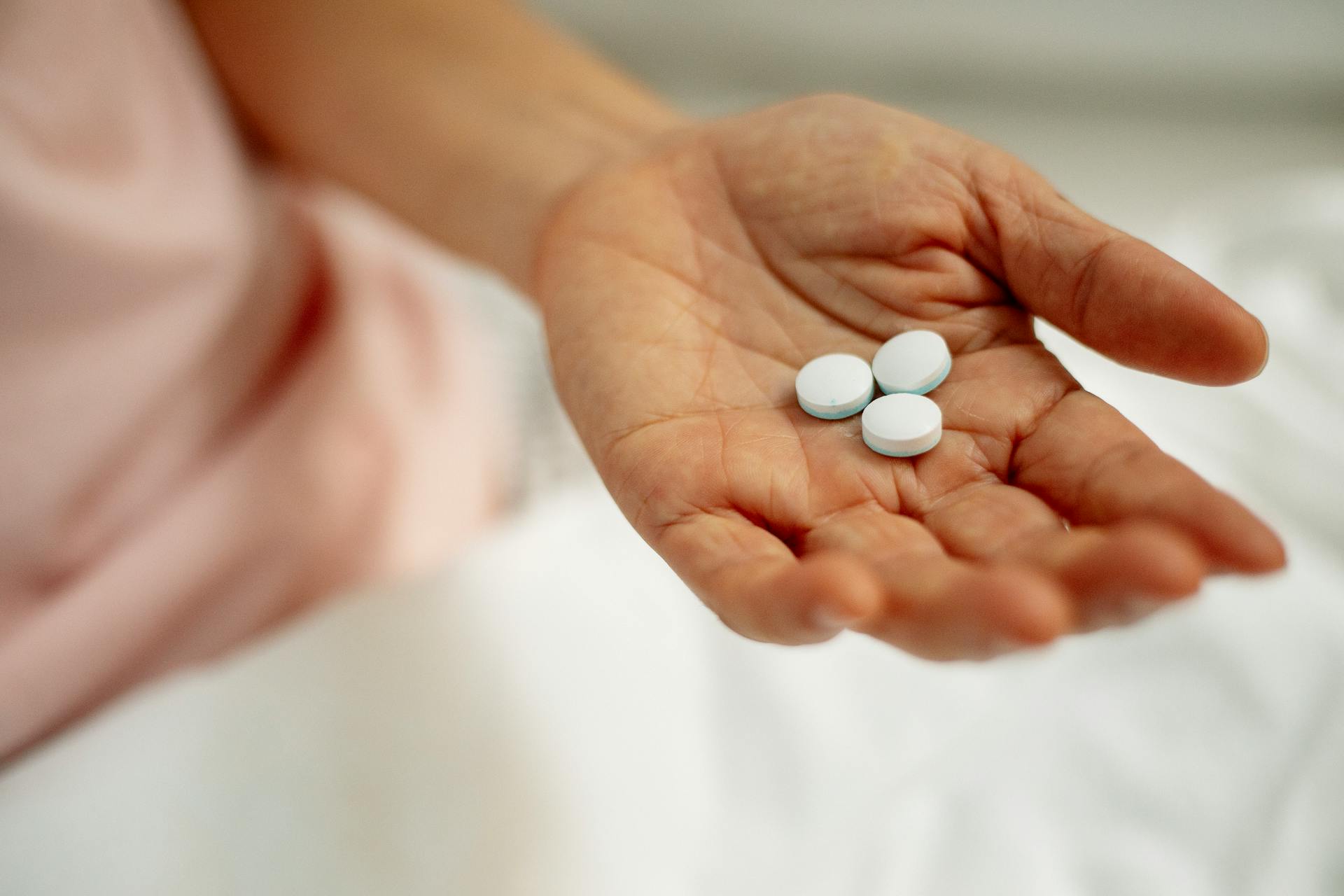 High angle of crop unrecognizable lady in sleepwear under blanket showing white medical pills in hand in light room