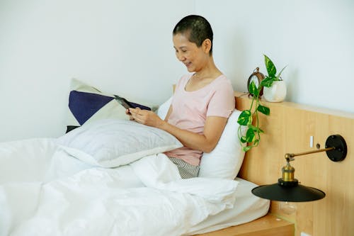 Positive middle age Asian lady in sleepwear surfing on mobile while sitting in bed with pillows under blanket next to potted green plant and lamp in light bedroom