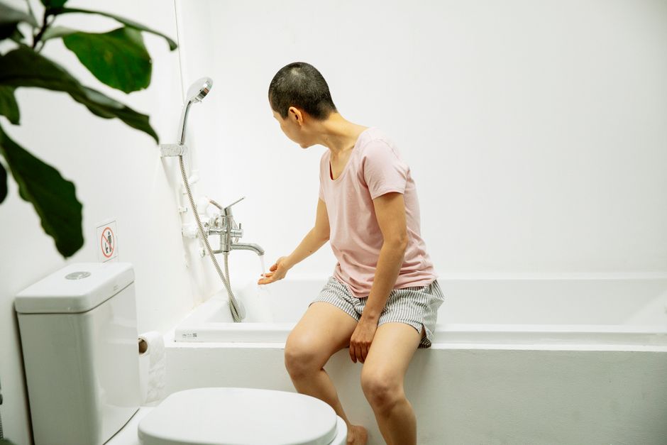 A woman sits on the bathtub edge in a clean, minimalist bathroom, touching the faucet.