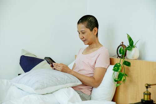Ethnic mature woman in pajama sitting in bed under blanket with pillows while using smartphone in bright bedroom near potted plant