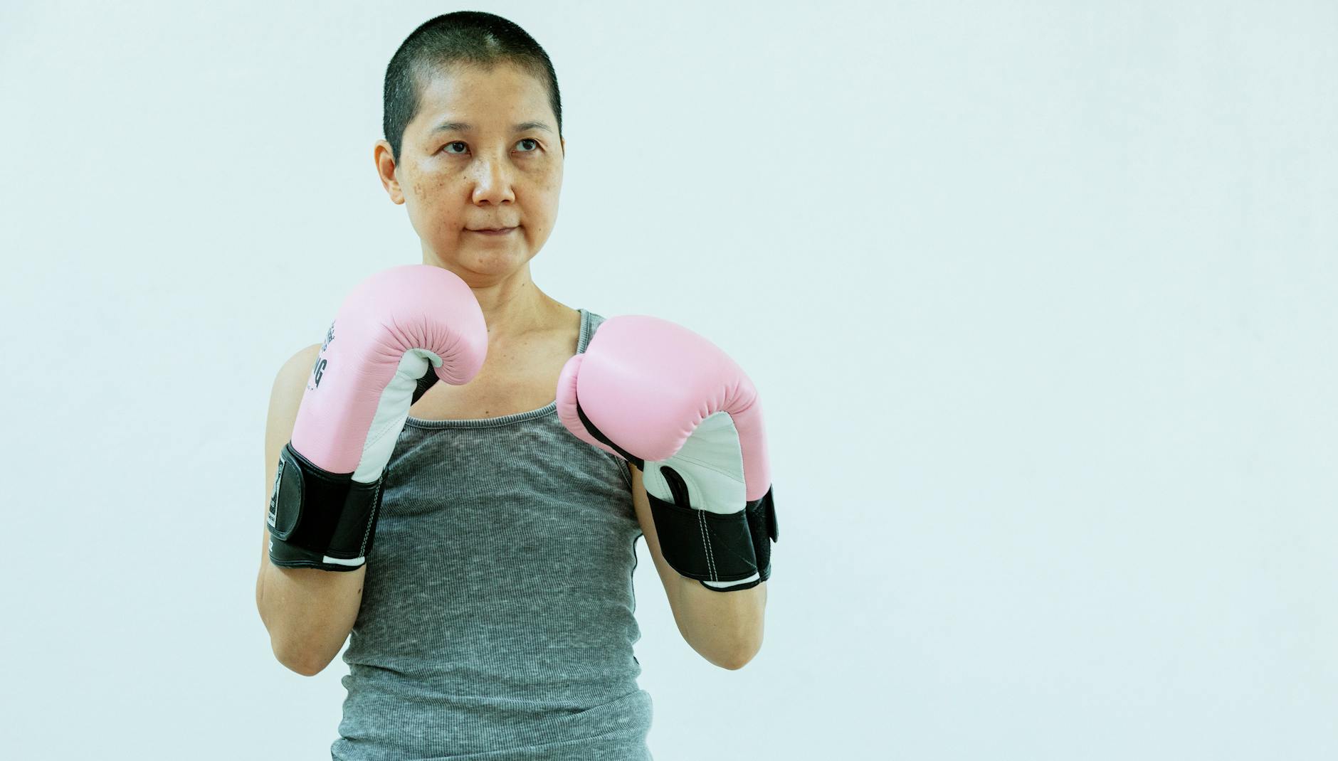Confident middle age ethnic woman wearing casual outfit and pink boxing gloves while preparing to fight on white background in light studio