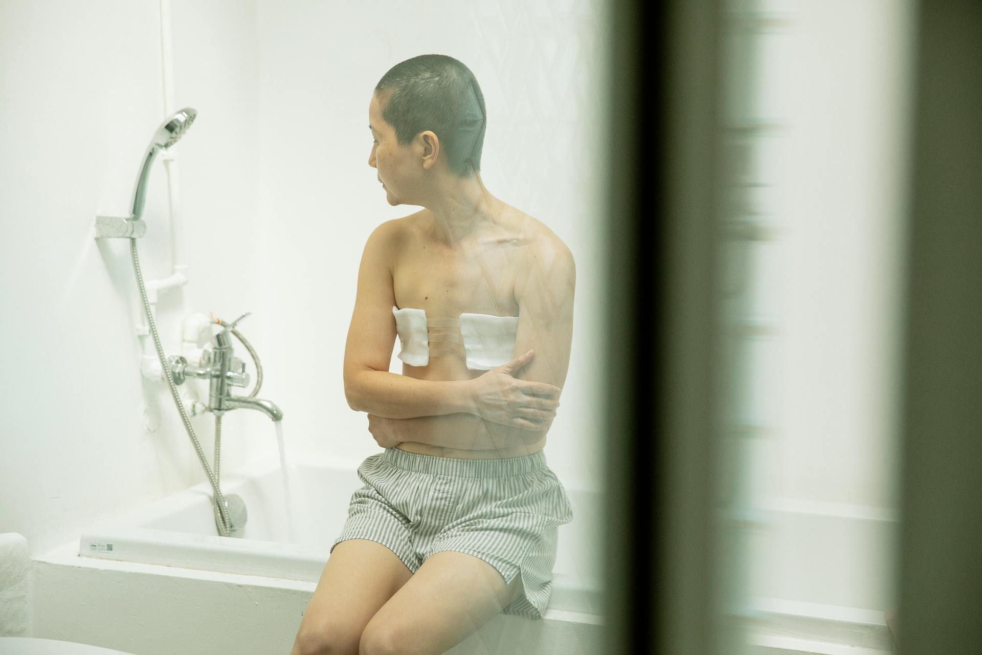 Through glass pensive shirtless woman with short dark hair and bandages on breast after surgery sitting on bath in bathroom at home