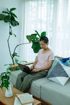 An Asian woman working on a laptop sitting on a modern sofa surrounded by plants in a bright home interior.