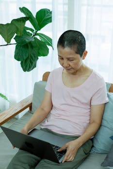 High angle of serious mature Asian lady in casual clothes sitting on couch while browsing on netbook next to green plant and window with curtains in light apartment