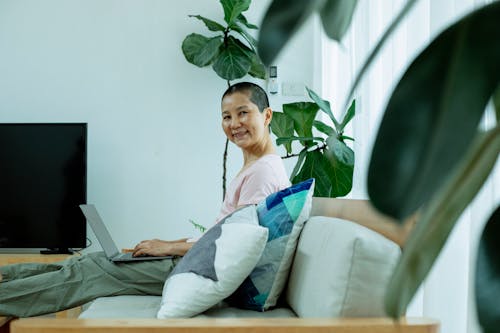 Side view of positive middle age ethnic female in casual outfit using laptop on sofa next to plants and window with curtains near TV in house and looking at camera