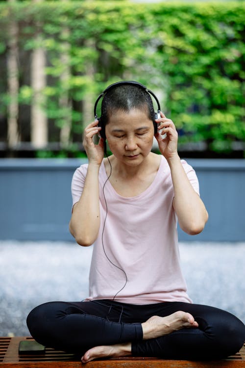 Full length of middle age Asian woman in casual outfit and headphones sitting with closed eyes and crossed legs on bench near phone in street near green plants in daytime