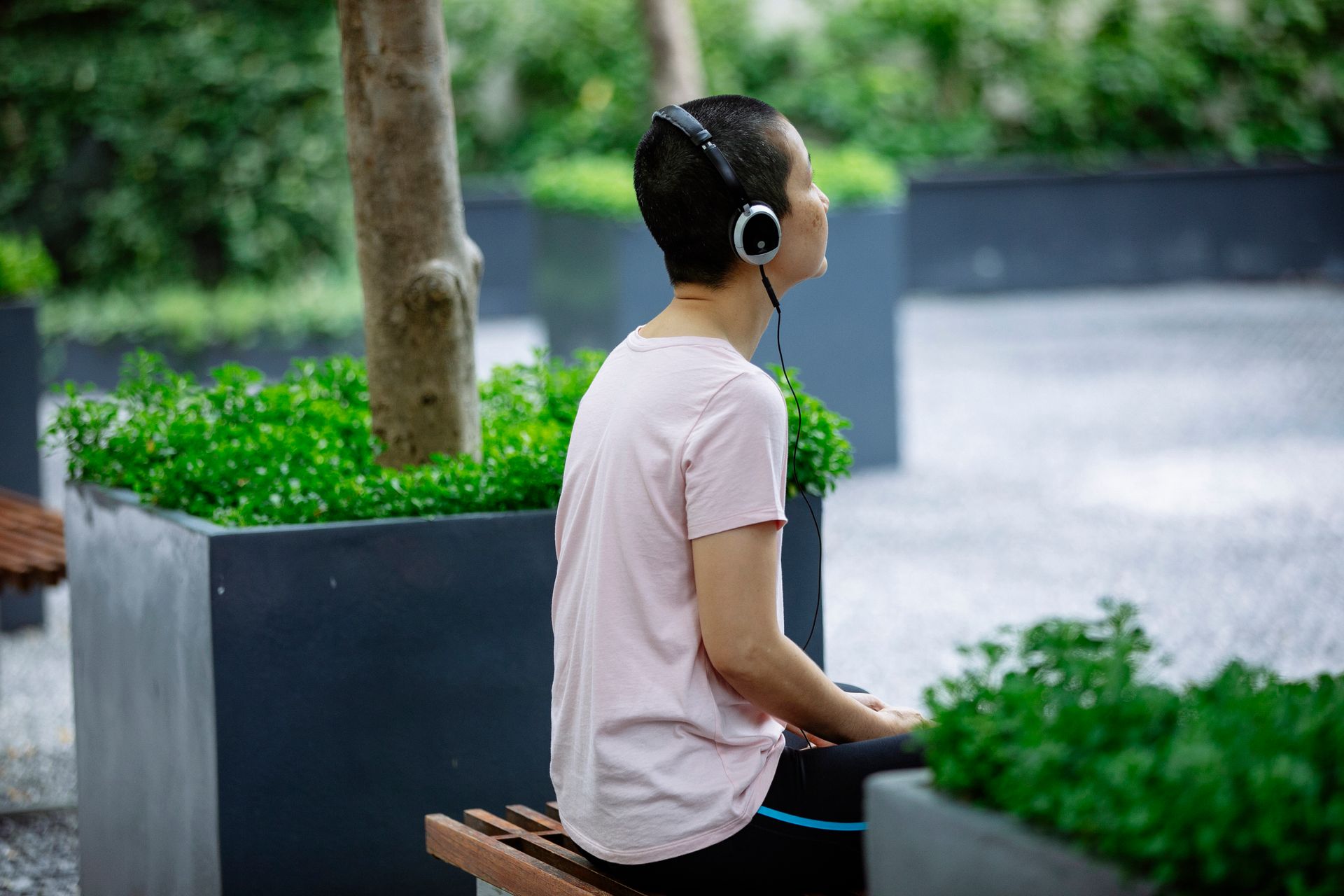 Faceless woman in casual outfit and headphones sitting on bench in street near green plants and trees in pots in summer day