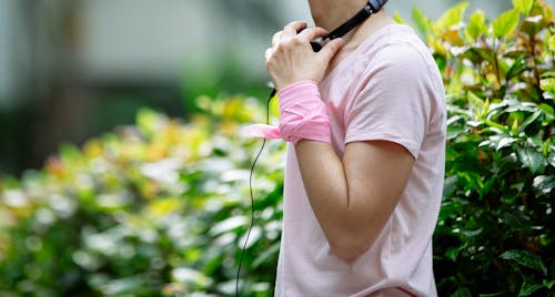 Crop faceless woman in casual clothes and pink bracelet with headphones standing near green bushes and plants with foliage in park in summer day