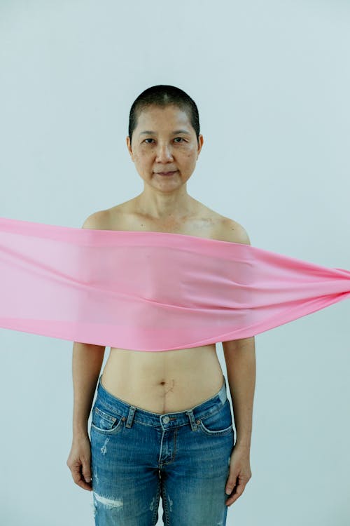 Slender ethnic female in jeans suffering from breast cancer and looking at camera on white background