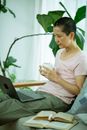 Tired ethnic female with cup of hot drink working with netbook near books at home with green plants