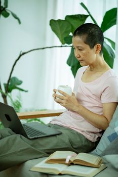 Tired ethnic female with cup of hot drink working with netbook near books at home with green plants