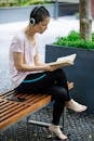 Serious ethnic woman in headphones reading novel on bench in park